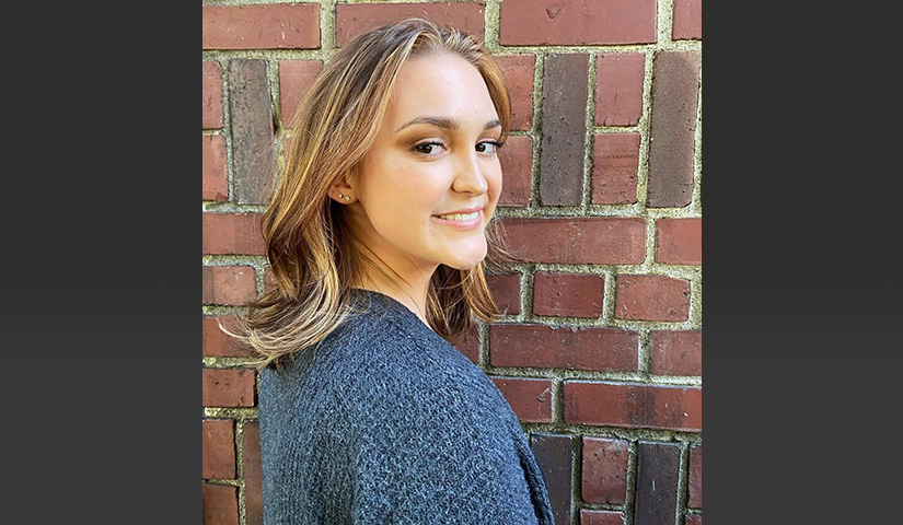 A portrait of research participant Amber wearing a gray-blue sweater and standing beside a brick wall