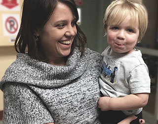 A mom holding and smiling at her son while the son looks at the camera and smiles