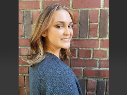 A portrait of research participant Amber wearing a gray-blue sweater and standing beside a brick wall